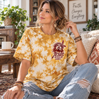 A woman sitting in a cozy living room wearing a mustard yellow and cream tie-dye t-shirt with a graphic of Jesus' face and the text 'JESUS I AM WITH YOU ALWAYS'.
