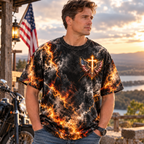 A man wearing a black graphic t-shirt with a fiery orange and yellow lightning pattern and a winged cross design, standing outdoors in front of a rustic wooden structure with an American flag and a motorcycle.