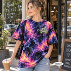 A woman wearing a vibrant purple, pink, and blue lightning-bolt tie-dye t-shirt with a 'JESUS CHRIST' graphic on the chest, standing in front of a storefront.