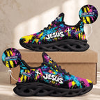 A pair of custom athletic sneakers with a vibrant multicolor graffiti-style paint splatter design in pink, yellow, blue, and purple on a black base. The side of the shoe features the text 'JESUS' in white with a gold cross and 'Custom Name' in white.
