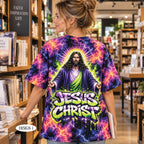 A woman standing in a bookstore, viewed from behind, wearing a colorful graphic t-shirt with a large print of Jesus Christ and the text 'JESUS CHRIST' on the back. The shirt features a purple, pink, and blue lightning tie-dye pattern.
