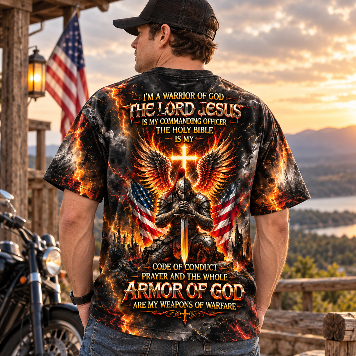 A man wearing a black baseball cap and blue jeans stands on a porch at sunset, shown from the back. He is wearing a short-sleeve graphic T-shirt with a large fiery design featuring a kneeling warrior, wings, American flags, and religious text.