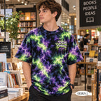 A man wearing a black t-shirt with a purple and green lightning bolt tie-dye pattern, standing in a bookstore. The shirt features a graphic on the left chest that reads 'JESUS CHRIST' in a stylized font.