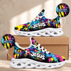 A pair of colorful sneakers displayed on a cardboard box against a beige wall. The shoes have a black base with bright neon paint-splatter graphics in pink, blue, yellow, and purple, plus white 'JESUS' text and cross graphics.