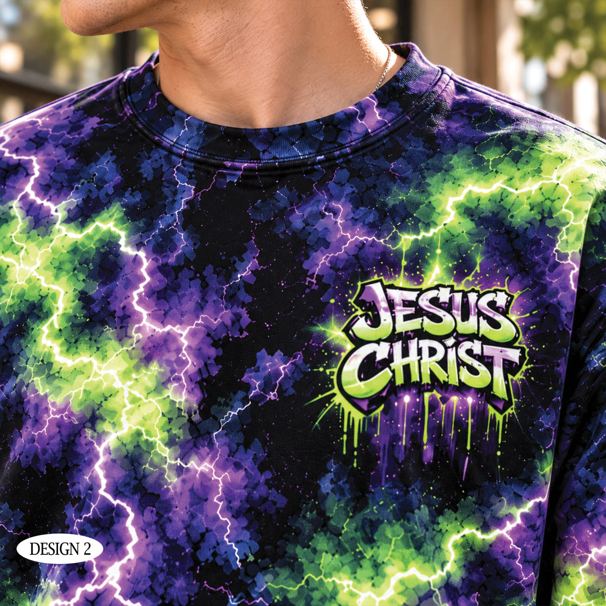 A close-up of a person wearing a short-sleeve crewneck t-shirt with a vibrant purple, green, and black lightning-bolt tie-dye pattern. The shirt features a prominent graphic on the chest that reads 'JESUS CHRIST' in a stylized, dripping graffiti font.