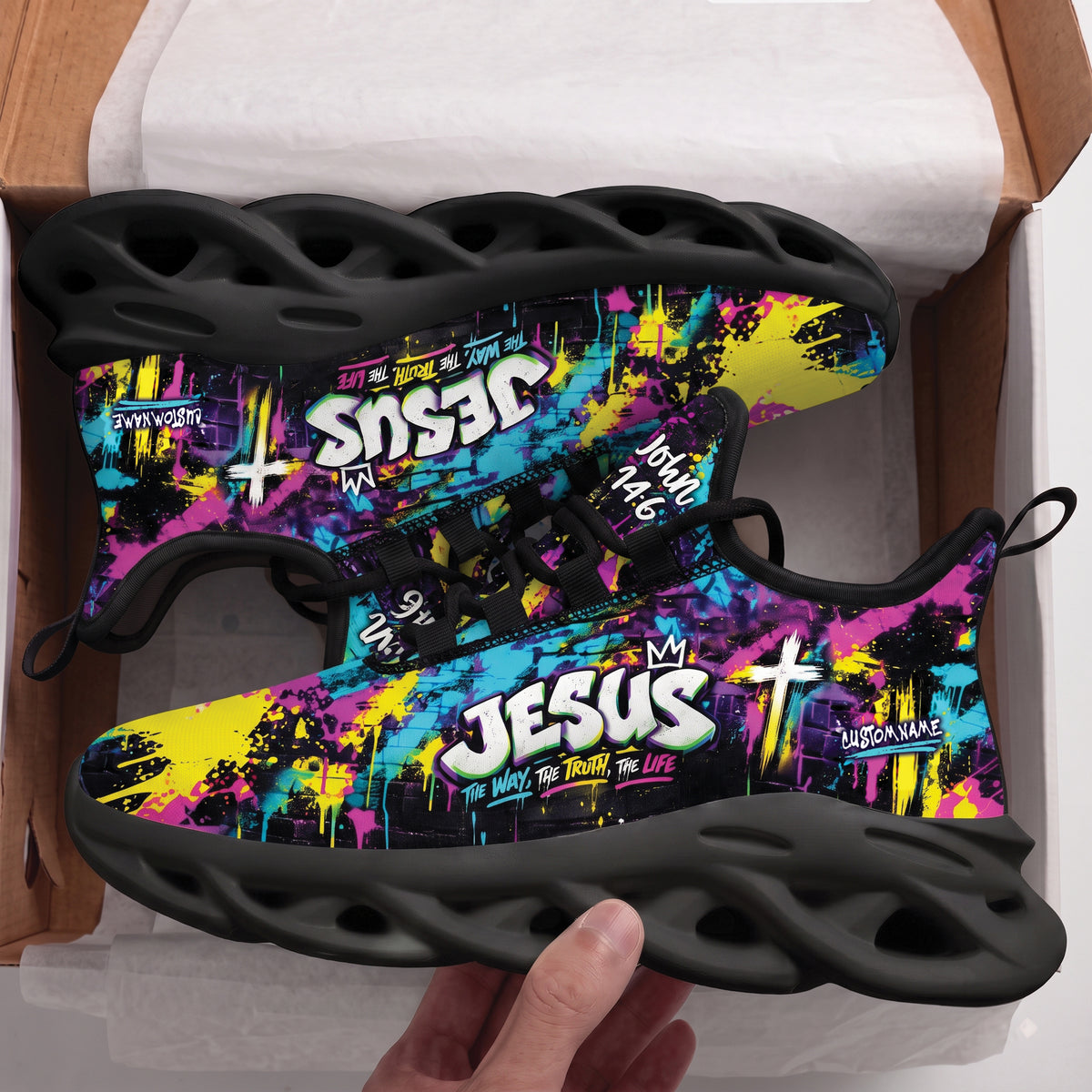 A pair of black athletic sneakers with a vibrant graffiti-style multicolor upper, held in a hand inside an open cardboard box. The shoes feature bold text including 'JESUS', 'John 14:6', and 'CUSTOM NAME' in white and yellow over a pink, blue, and yellow background.