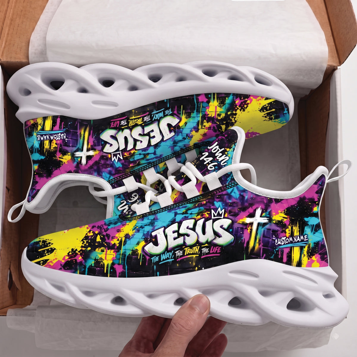 A pair of custom sneakers with a colorful graffiti-style print featuring the text 'JESUS' and 'CUSTOM NAME', displayed inside a shoebox with a hand holding one shoe.